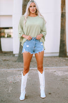 Woman wearing sage Lines Drawn oversized top with denim shorts and white boots, showcasing casual street style.