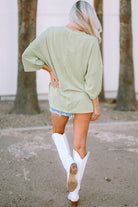 Woman wearing sage Lines Drawn Top with roll-tab sleeves and white boots, showcasing casual outdoor fashion style.