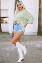 Sage green oversized ribbed knit top with roll-tab sleeves, paired with denim shorts and white boots, styled for a chic casual look.