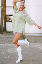 Woman wearing an oversized sage Lines Drawn Top with roll-tab sleeves, paired with denim shorts and white boots, showcasing textured style.