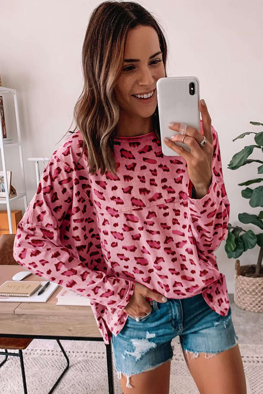 Woman wearing pink leopard print long sleeve top with relaxed fit, styled with denim shorts and smiling in a cozy setting.