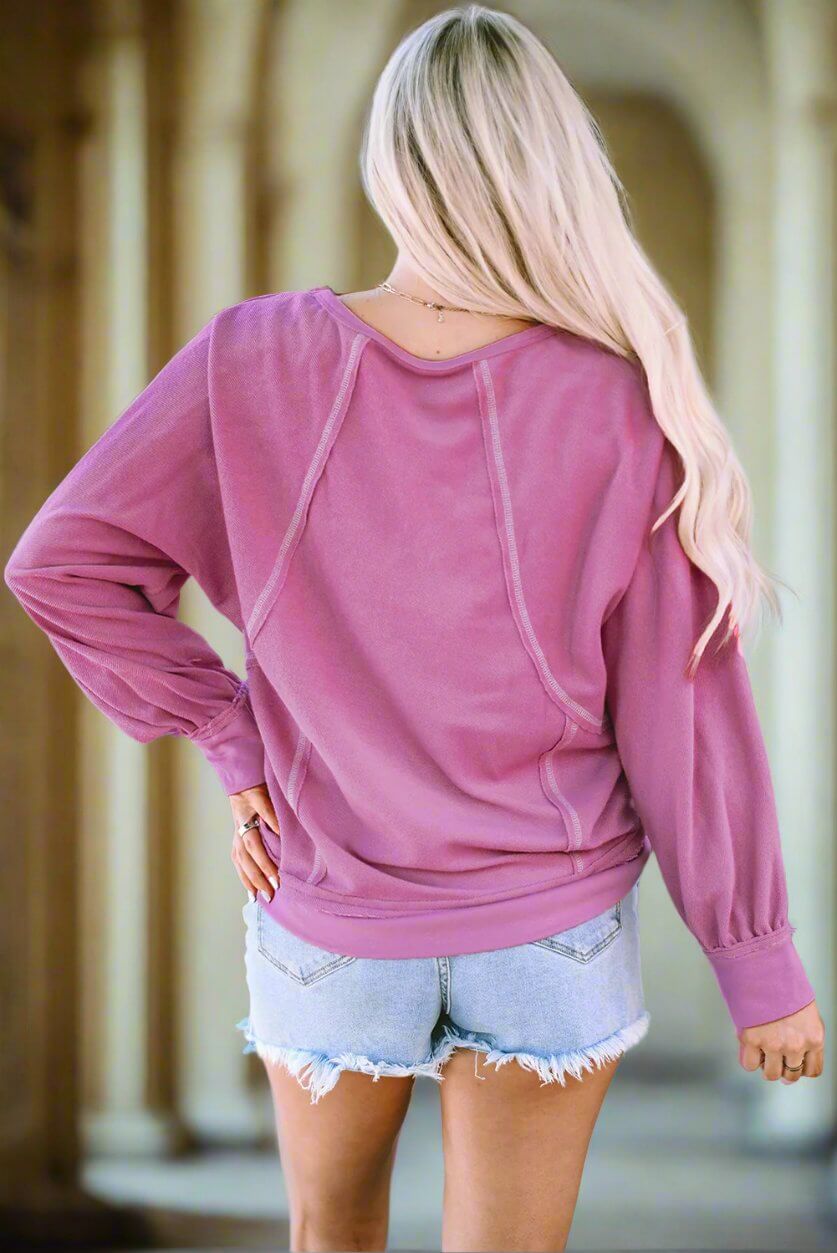 Back view of a woman in a pink "BEAUTY IN MOTION" top with exposed seam details, paired with denim shorts.