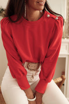 Woman wearing a red top with Bishop sleeves and button-tab detail, paired with white pants, sitting in a cozy room.