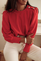 Woman in a stylish red top with Bishop sleeves and button-tab detail, perfect for fall fashion, paired with white pants.