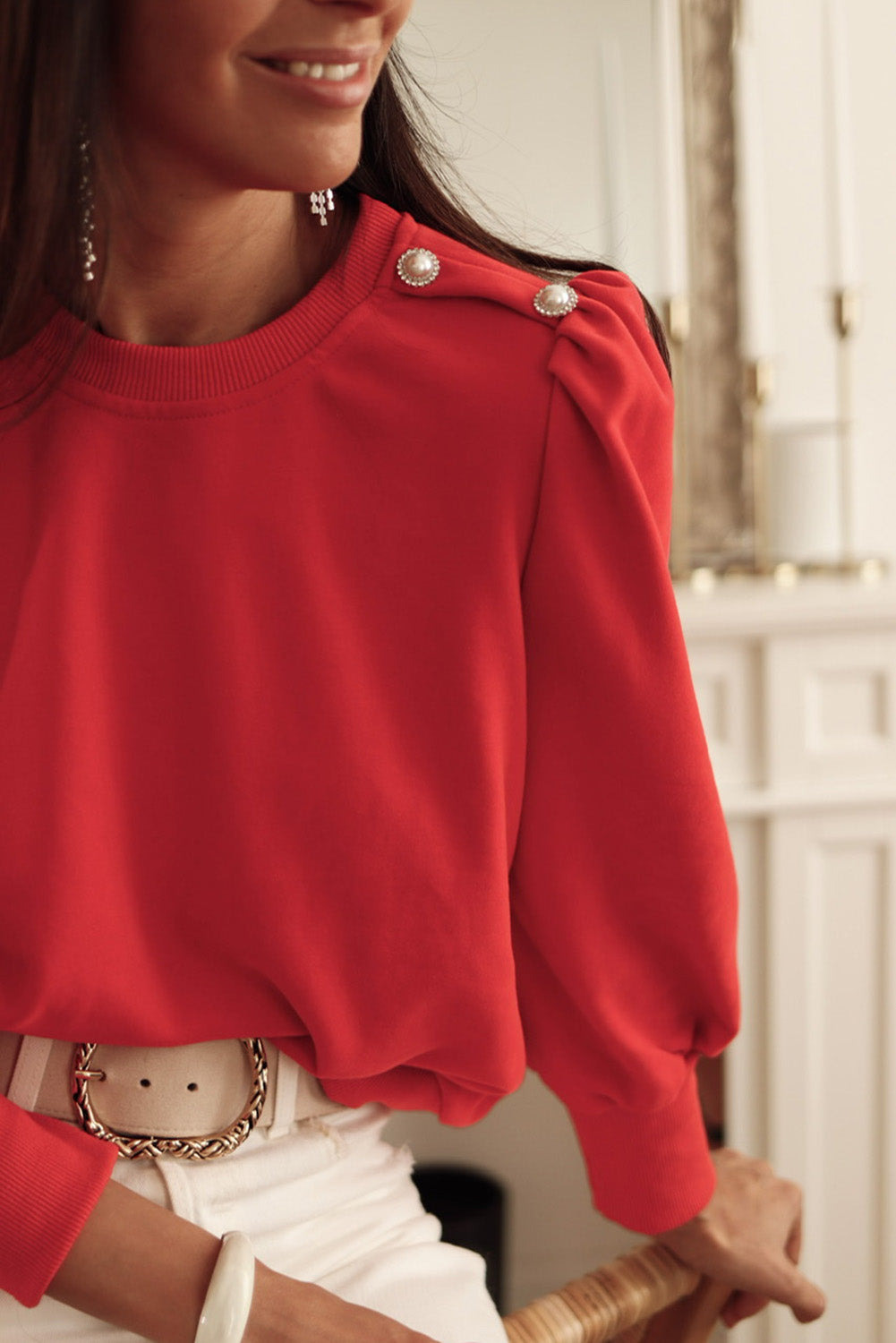 Woman wearing red top with elegant Bishop sleeves and button-tab detail, accessorized with a white belt and jewelry, smiling indoors.