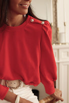 Woman wearing red top with elegant Bishop sleeves and button-tab detail, accessorized with a white belt and jewelry, smiling indoors.