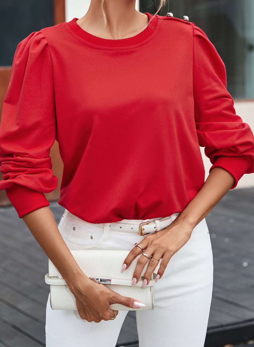 Woman wearing a stylish red top with Bishop sleeves and button-tab detail, paired with white pants and a white clutch.