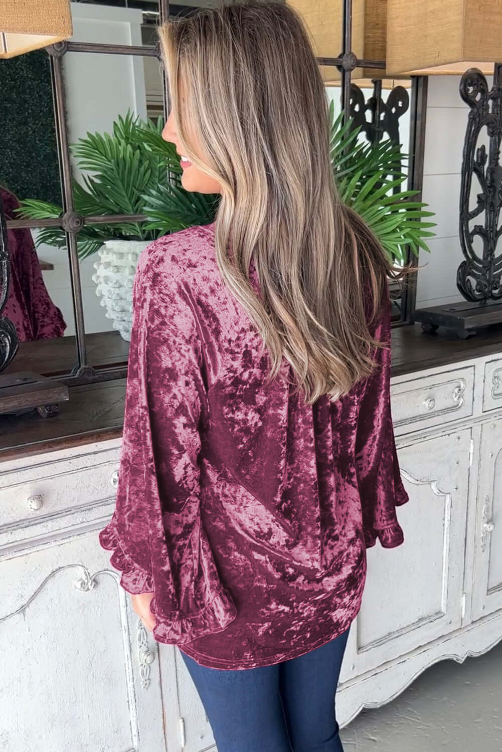 Woman wearing Velvet Whisper blouse in rose pink with flowy ruffled sleeves, standing in front of white vintage dresser.