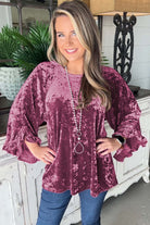 Woman wearing Velvet Whisper blouse in rose pink with ruffled pagoda sleeves and necklace, posing indoors.