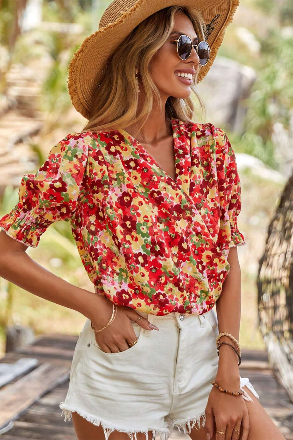 Model wearing Tropical Tango red floral blouse with bubble sleeves and shorts, enjoying a sunny outdoor setting.