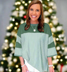 Woman smiling in a green striped patchwork top, standing in front of a festive tree, showcasing a relaxed fit and cozy style.