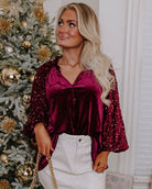 Model wearing a sequined sleeve velvet top with ruffle details, styled for a holiday occasion by a decorated Christmas tree.