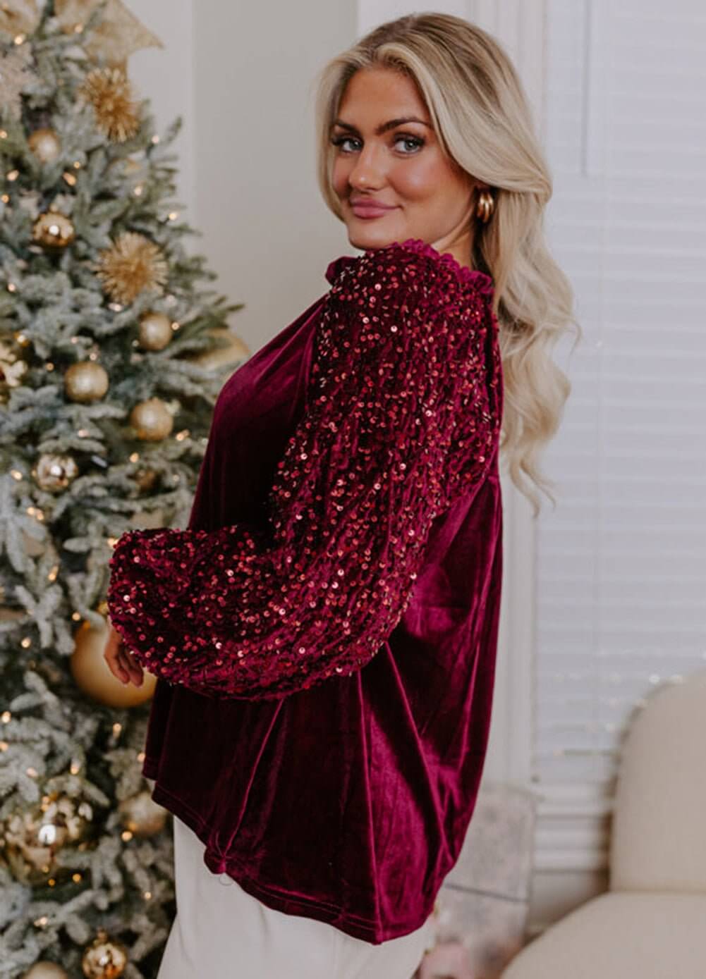 Woman models SEQUIN SIREN velvet top with sequined sleeves, posing by a holiday tree, showcasing glamour and style.