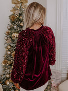 Woman in a velvet SEQUIN SIREN top with ruffled neckline and sequined sleeves, posing near a decorated Christmas tree.