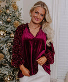 Model wearing SEQUIN SIREN velvet top with sequined sleeves, posing by a decorated holiday tree. Perfect for festive occasions.