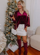 Woman wearing a stunning sequin-accented velvet top, white skirt, and knee-high boots, celebrating by a festive Christmas tree.