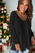 Woman smiling in a black waffle knit top with V-neckline, standing in front of a festive Christmas tree.