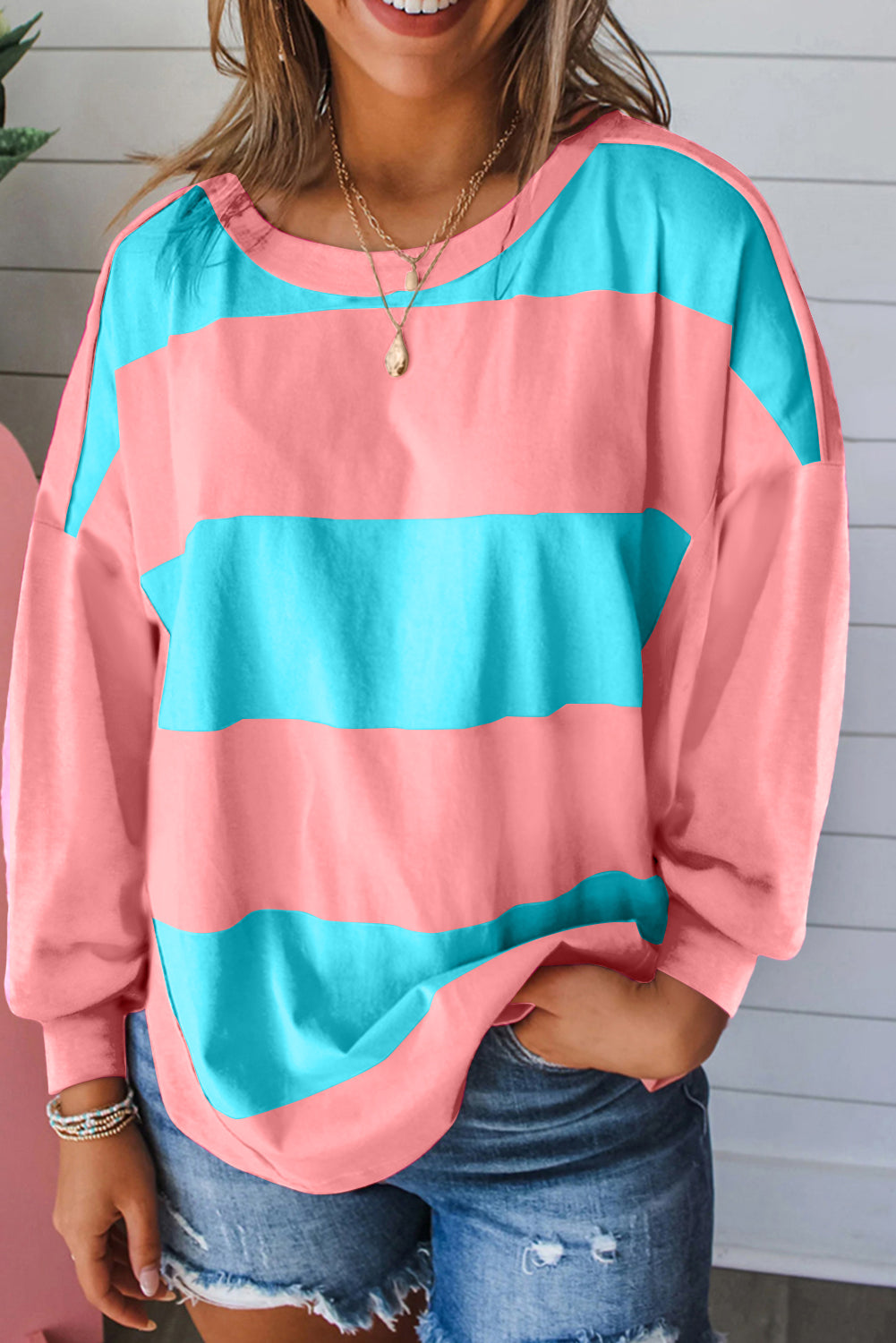 Woman wearing a blue and pink striped crew neck top with drop shoulders and vibrant color block design, paired with denim shorts.