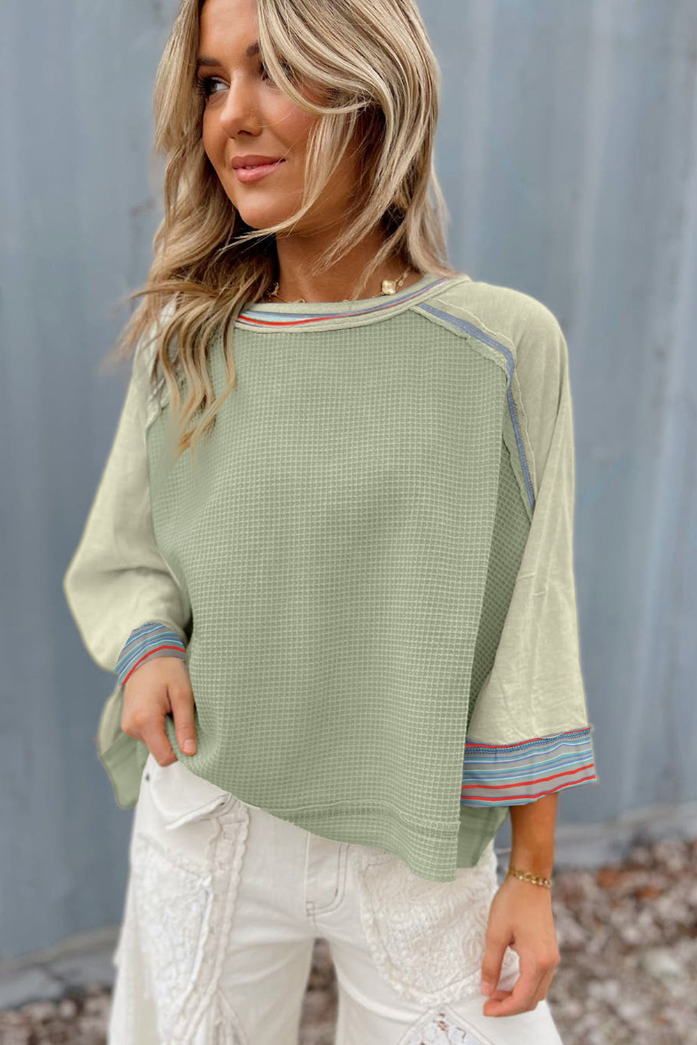 Woman wearing green waffle knit top with wide bracelet sleeves, contrast stripe stitching, and raw hem, paired with white pants.