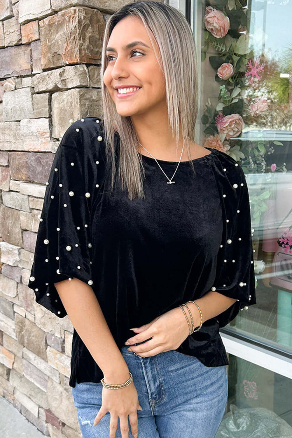 Model in black velvet MIDNIGHT PEARLS TOP with pearl embellishments and half puff sleeves, smiling outdoors.