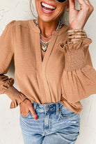 Stylish woman in a tan V-neck blouse with shirred cuffs, paired with denim, smiling and accessorized with layered necklaces.