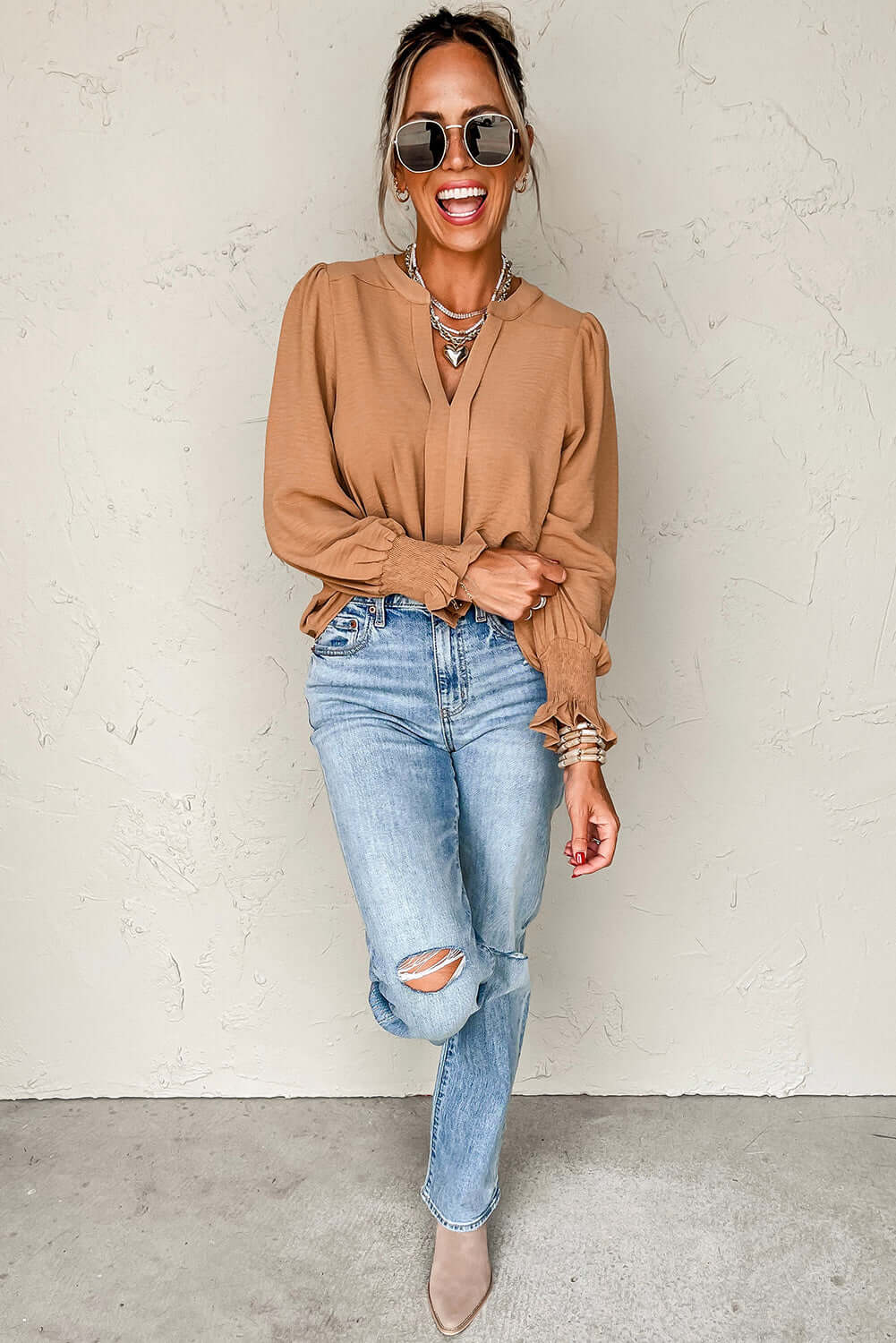 Woman wearing a beautiful beige V-neck blouse with shirred cuffs paired with ripped jeans and sunglasses, smiling against a neutral background.