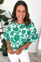 Woman smiling in a green floral Jade Blooms Top with ruffled sleeves, paired with white pants.