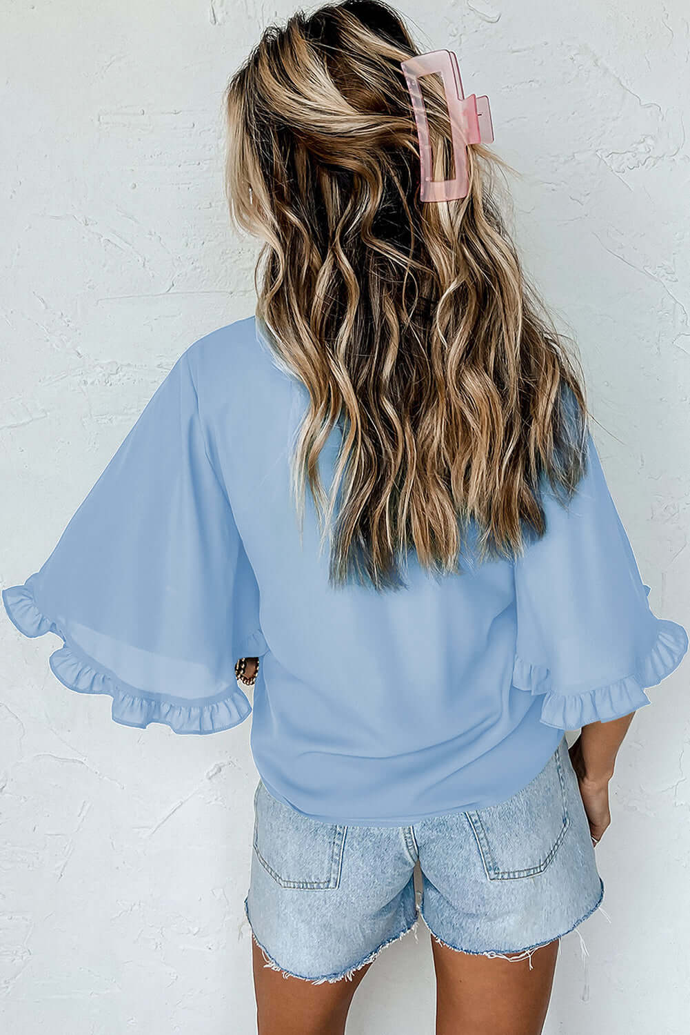 Back view of a woman wearing a sky blue Frill Seeker Top with ruffle sleeves and denim shorts, featuring stylish hair accessory.
