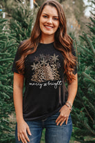 Woman wearing a Merry & Bright Leopard Tee with Christmas tree graphic, standing among trees, showcasing holiday style.