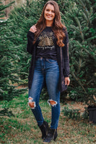 Woman wearing Merry & Bright Leopard Tee with Christmas tree design, paired with jeans and a dark cardigan, standing among trees.