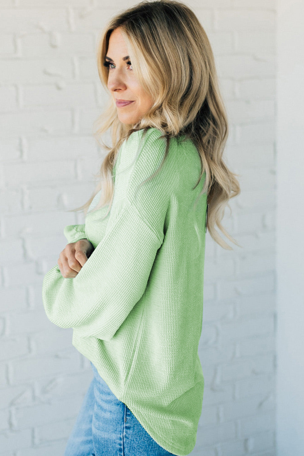 Woman wearing mint-colored top with corded ribbing, drop shoulder design, and relaxed fit pairing with denim jeans against brick wall.