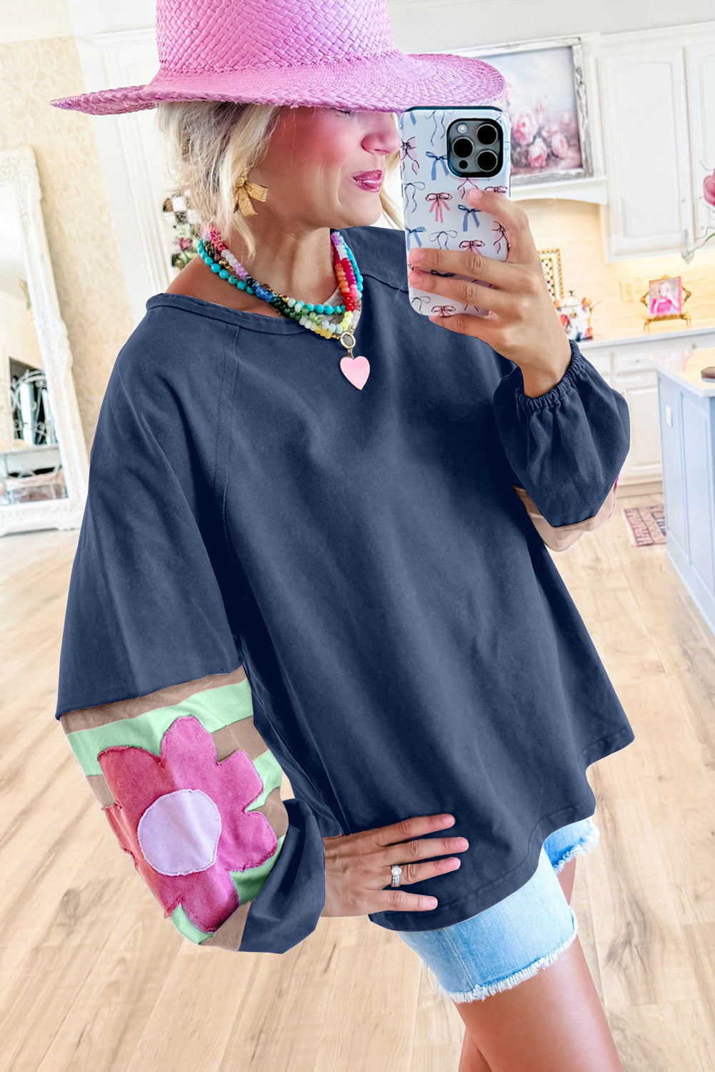 Woman wearing blue oversized pullover with floral sleeves, paired with a pink hat and colorful necklace in a stylish room.