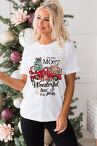 Woman wearing "Most Wonderful Time" T-shirt with red truck design, standing by a decorated Christmas tree, showcasing holiday spirit.