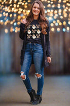 Woman wearing festive floral sweater and jeans, standing in front of blurred string lights backdrop