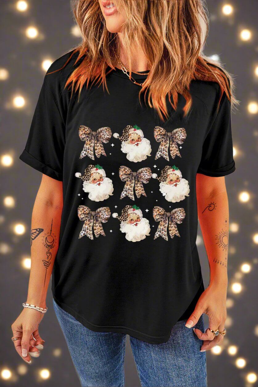 Festive black T-shirt with leopard print bows and Santa Claus graphics, modeled with jeans in front of a holiday lights background.