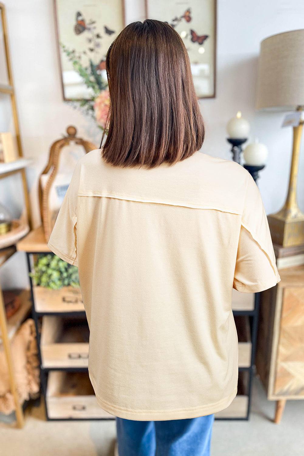 Back view of a woman in a vanilla floral patch top with exposed stitching, styled in a cozy boutique setting.