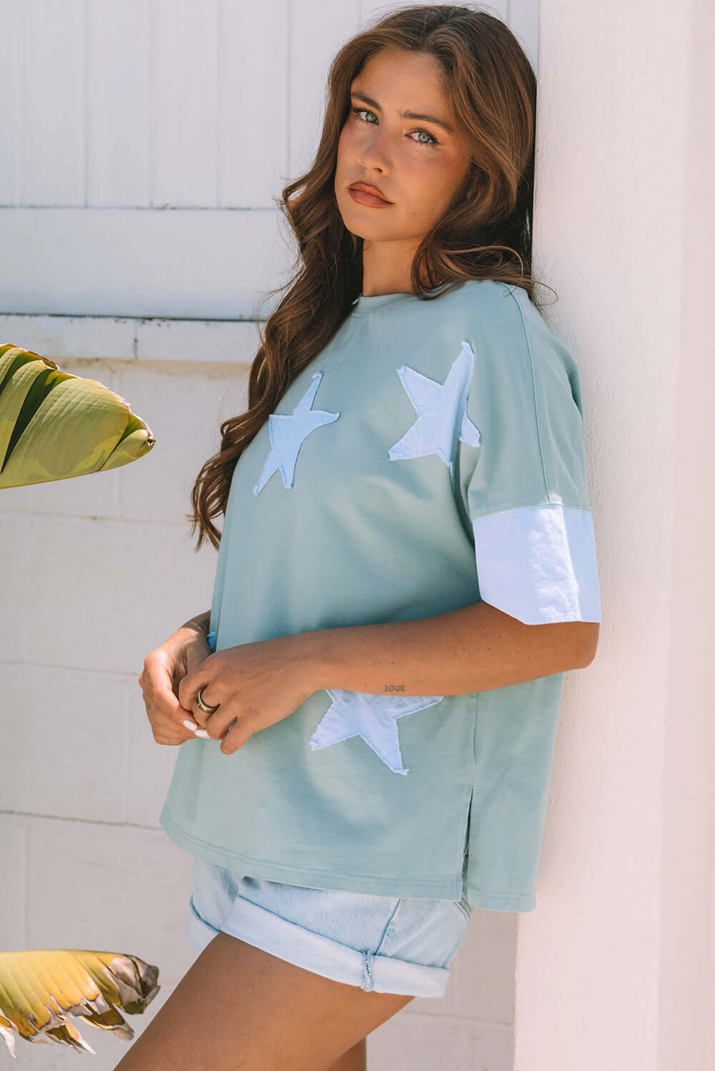 Oversized sage blue Cosmic Crush top with sky star patches, paired with denim shorts in a casual outdoor setting.