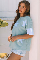 Oversized sage blue Cosmic Crush top with sky star patches, paired with denim shorts in a casual outdoor setting.