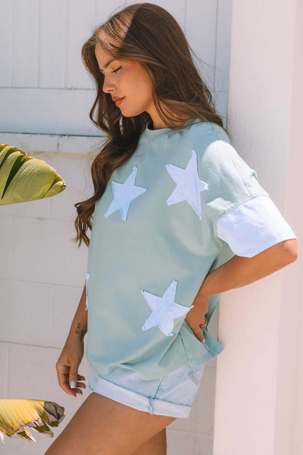 Woman in stylish sage oversized tee with sky blue star patches, posing against a white wall.