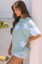 Woman in stylish sage oversized tee with sky blue star patches, posing against a white wall.