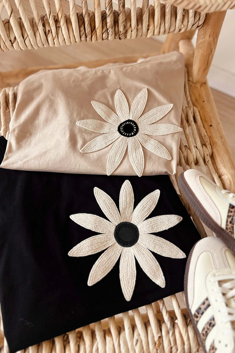 Two tees featuring crochet daisy graphics, one beige and one black, displayed with stylish sneakers on a woven surface.