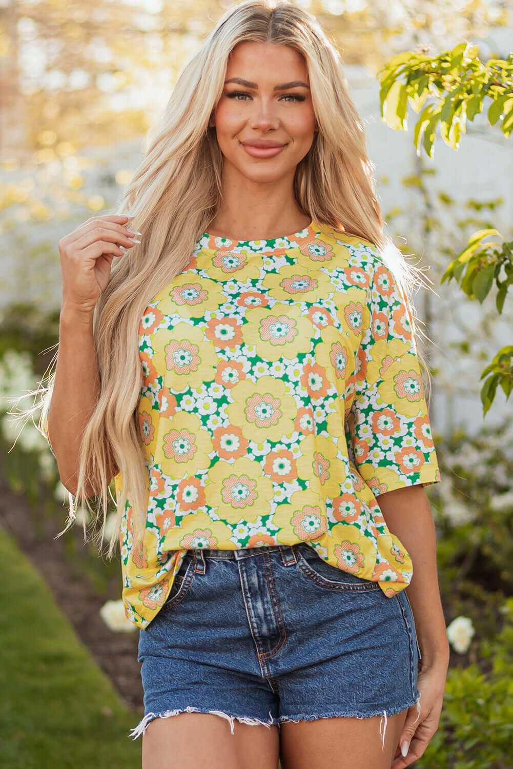 Model wearing vibrant yellow Daisy Disco Top with floral print, paired with denim shorts in a sunlit garden.