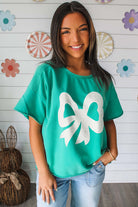 Teal lace bow tee, Ribbon Theory Top, casual style, playful design, model smiling in a bright room.