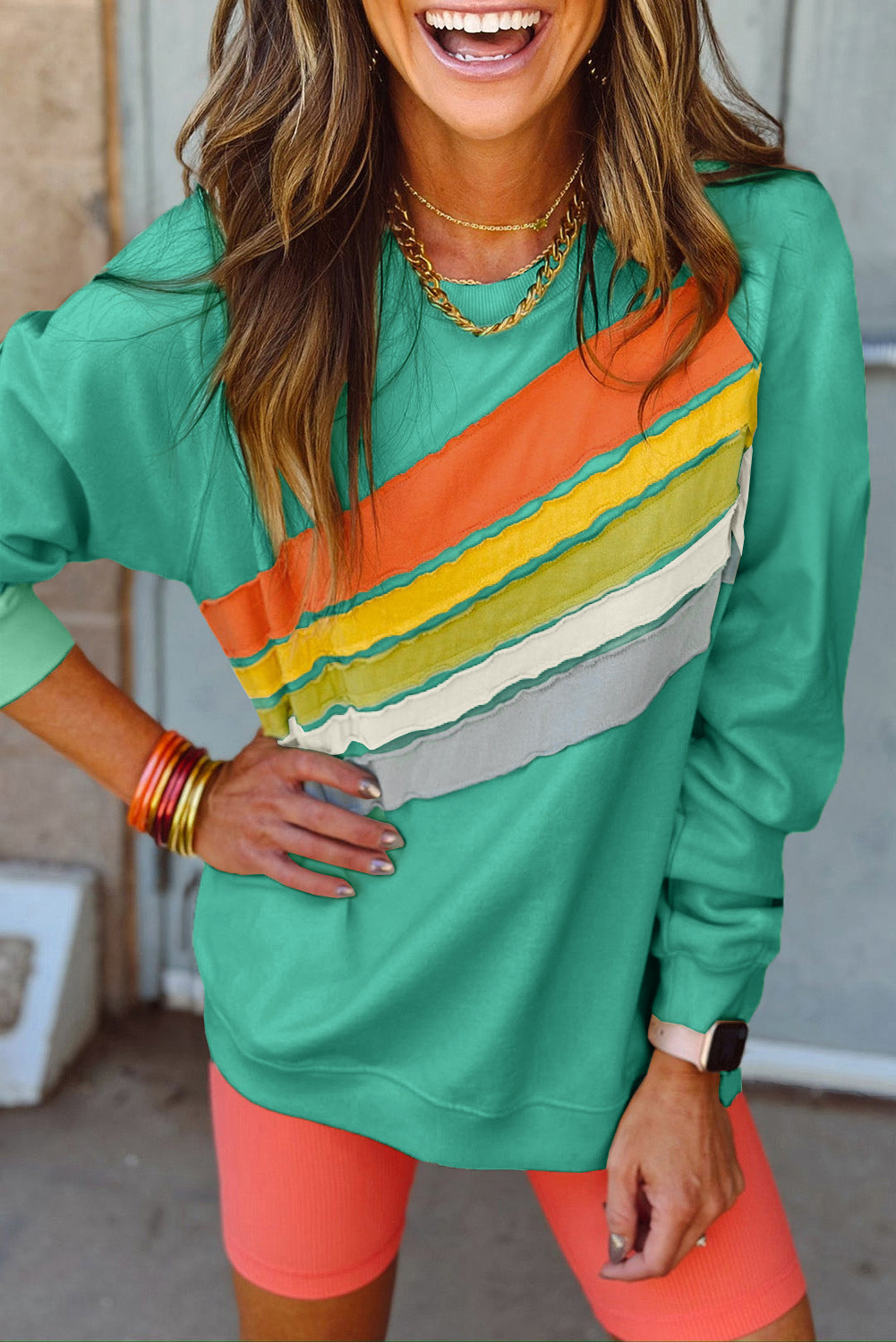 Woman wearing Upward Trends aqua pullover with rainbow color-block design, paired with orange shorts and gold jewelry, smiling outdoors.