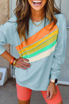 Woman wearing sky blue crewneck with rainbow color-block design and orange shorts, accessorized with bracelets and watch.