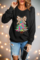 Woman wearing black sweatshirt with colorful Christmas tree and animal-print bow, paired with jeans, holding a tumbler.