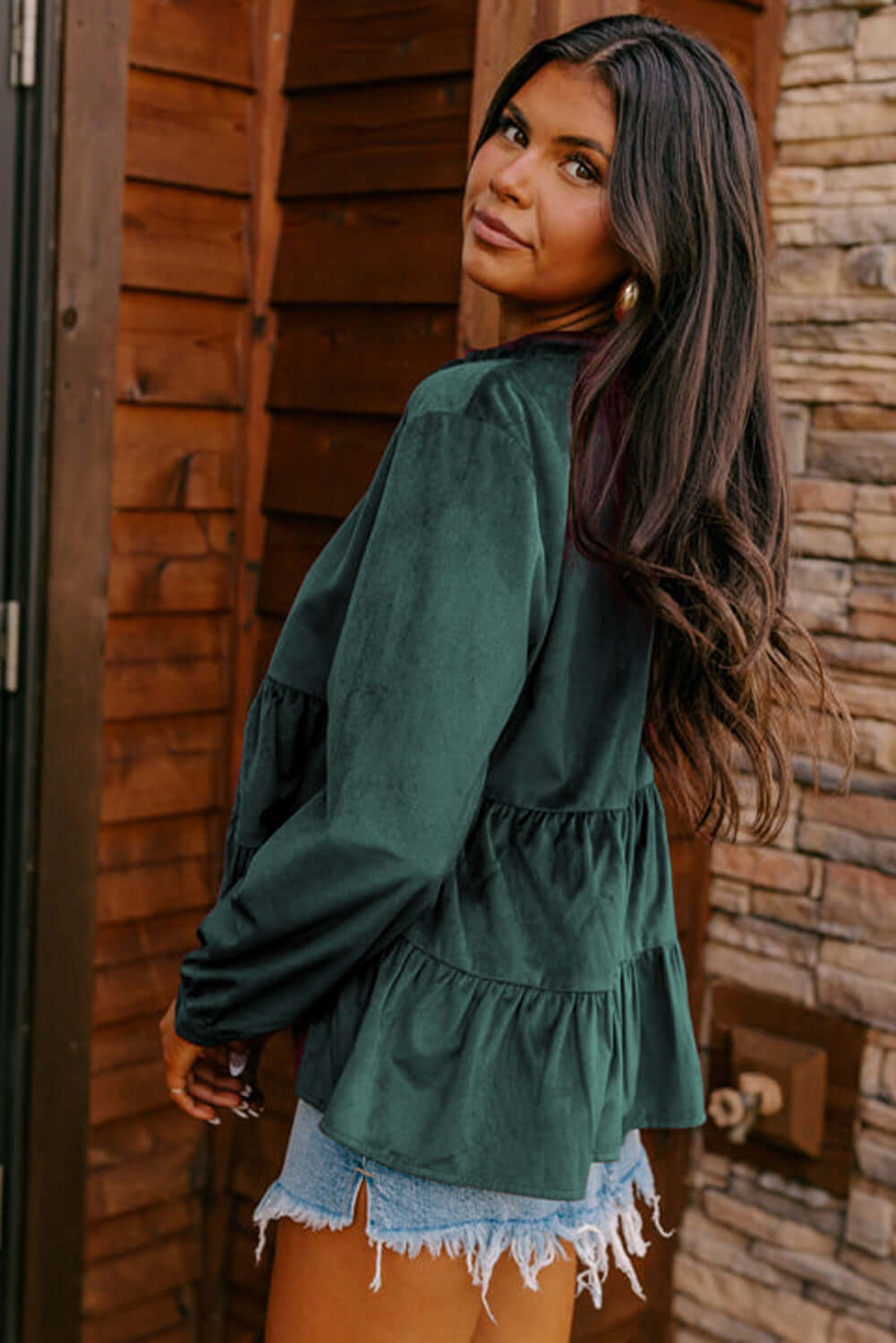 Woman wearing State of Grace top in forest green with ruffle tiers and O neck, paired with denim shorts, standing by a wooden wall.