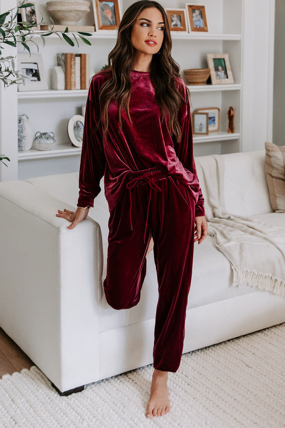 Woman wearing burgundy velvet two-piece set with long sleeve top and drawstring pants, sitting on a white sofa in a cozy room.