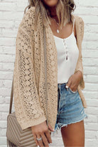 Woman wearing a cable knit apricot beige cardigan with a white top and denim shorts against a white brick wall.