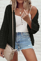Model wearing the Café Date Cardigan in jet black, styled with a white top and denim shorts against a brick background.
